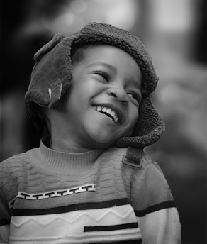 A young child with a joyful expression, wearing a warm, patterned sweater and a cozy hat. The child is looking upwards, smiling broadly, and appears to be in a moment of laughter.