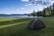 A serene lakeside scene with a grassy area in the foreground. A tent is pitched on the grass, and two picnic tables are scattered nearby. The lake is calm, reflecting the blue sky dotted with clouds. In the distance, hills and a small boathouse are visible across the water. Trees frame the scene, adding to the tranquil, natural setting.
