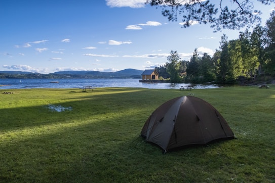 A serene lakeside scene with a grassy area in the foreground. A tent is pitched on the grass, and two picnic tables are scattered nearby. The lake is calm, reflecting the blue sky dotted with clouds. In the distance, hills and a small boathouse are visible across the water. Trees frame the scene, adding to the tranquil, natural setting.