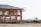A wooden house frame under construction with scaffolding surrounding it. The setting appears to be a construction site with dirt ground and some fencing. In the background, there are open fields and distant trees.