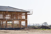 A wooden house frame under construction with scaffolding surrounding it. The setting appears to be a construction site with dirt ground and some fencing. In the background, there are open fields and distant trees.