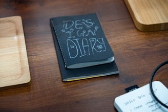 A black notebook titled 'Design Diary' is placed on a wooden table, flanked by two wooden trays. In the corner, there is a white notepad with handwritten notes visible, and a black cable is slightly coiled on the right side of the image.