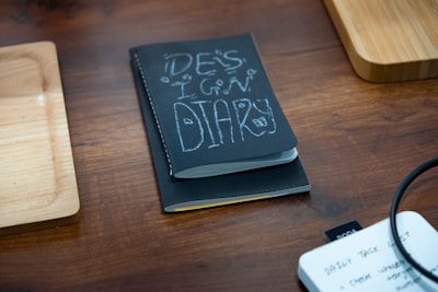 A black notebook titled 'Design Diary' is placed on a wooden table, flanked by two wooden trays. In the corner, there is a white notepad with handwritten notes visible, and a black cable is slightly coiled on the right side of the image.