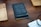 A black notebook titled 'Design Diary' is placed on a wooden table, flanked by two wooden trays. In the corner, there is a white notepad with handwritten notes visible, and a black cable is slightly coiled on the right side of the image.