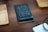 A black notebook titled 'Design Diary' is placed on a wooden table, flanked by two wooden trays. In the corner, there is a white notepad with handwritten notes visible, and a black cable is slightly coiled on the right side of the image.
