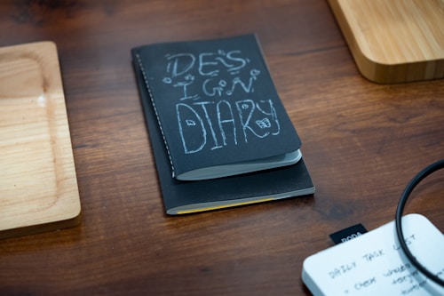 A black notebook titled 'Design Diary' is placed on a wooden table, flanked by two wooden trays. In the corner, there is a white notepad with handwritten notes visible, and a black cable is slightly coiled on the right side of the image.