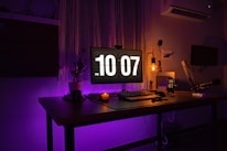 A darkly lit workspace featuring a large monitor displaying a digital clock reading 10:07 PM. The desk has a keyboard, a mouse, a candle, and small plant pots. There's an industrial-style lamp providing a warm glow. The background includes a curtain, an air conditioner, and a wall-mounted microphone.