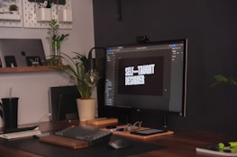 A workspace with a modern setup including a computer monitor displaying the words 'Self-taught designer'. The desk features a keyboard, mouse, glasses, and a potted plant. An open laptop is visible on a wooden shelf along with other small items. The area has a neat and organized appearance with a plant providing a touch of greenery.