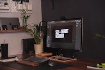 A workspace with a modern setup including a computer monitor displaying the words 'Self-taught designer'. The desk features a keyboard, mouse, glasses, and a potted plant. An open laptop is visible on a wooden shelf along with other small items. The area has a neat and organized appearance with a plant providing a touch of greenery.