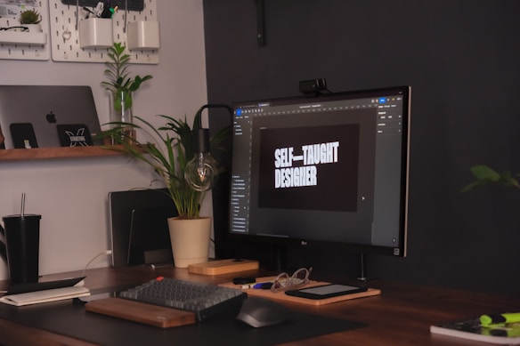 A workspace with a modern setup including a computer monitor displaying the words 'Self-taught designer'. The desk features a keyboard, mouse, glasses, and a potted plant. An open laptop is visible on a wooden shelf along with other small items. The area has a neat and organized appearance with a plant providing a touch of greenery.