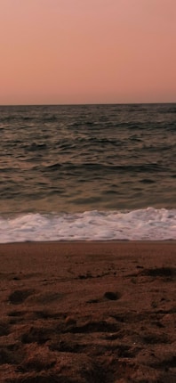 A serene beach scene at sunset with gentle waves lapping at the sandy shore. The sky is painted in soft hues of pink and orange, creating a calm and peaceful atmosphere.