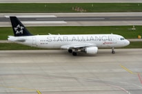 An airplane with the text STAR ALLIANCE on its body stands on the tarmac. The aircraft is mostly white with a black tail featuring a star logo. The red and white SWISS logo is displayed near the front. The surroundings include a runway and grass areas.