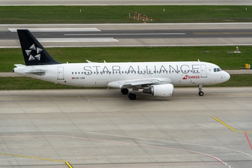 An airplane with the text STAR ALLIANCE on its body stands on the tarmac. The aircraft is mostly white with a black tail featuring a star logo. The red and white SWISS logo is displayed near the front. The surroundings include a runway and grass areas.