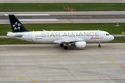 An airplane with the text STAR ALLIANCE on its body stands on the tarmac. The aircraft is mostly white with a black tail featuring a star logo. The red and white SWISS logo is displayed near the front. The surroundings include a runway and grass areas.