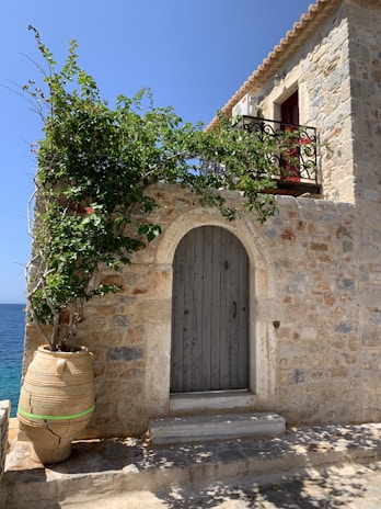 A charming Mediterranean-style stone house with a large clay pot containing a leafy plant. The blue sea can be seen in the background. A wooden door is set into the stone wall next to a small staircase, and a wrought iron balcony with greenery adds to the rustic appeal.