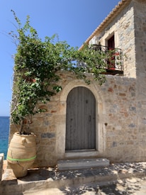 A charming Mediterranean-style stone house with a large clay pot containing a leafy plant. The blue sea can be seen in the background. A wooden door is set into the stone wall next to a small staircase, and a wrought iron balcony with greenery adds to the rustic appeal.