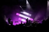 A concert stage with silhouettes of musicians performing, including a guitarist, drummer, and keyboard player. The background features a large display of pink and white geometric shapes, and the stage is illuminated with purple lighting, creating a vibrant and dynamic atmosphere.