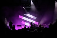 A concert stage with silhouettes of musicians performing, including a guitarist, drummer, and keyboard player. The background features a large display of pink and white geometric shapes, and the stage is illuminated with purple lighting, creating a vibrant and dynamic atmosphere.