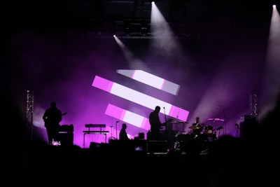 A concert stage with silhouettes of musicians performing, including a guitarist, drummer, and keyboard player. The background features a large display of pink and white geometric shapes, and the stage is illuminated with purple lighting, creating a vibrant and dynamic atmosphere.