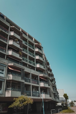 A multi-story residential building with a modern, concrete exterior featuring several balconies and windows. The building is angled in the photo to emphasize its height and geometric structure. A few trees are visible in the foreground, adding greenery to the urban setting.