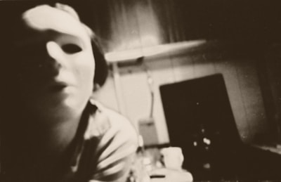 A person wearing a white, expressionless mask in a dimly lit room. The background is blurred, showing indistinct furniture and objects, with a sepia-toned color dominating the image.