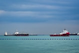Large ships are positioned on a calm ocean under a cloudy sky. The sea appears serene with visible waves, and there is a line of blue barriers floating on the water. The ships vary in size and are distributed across the horizon, indicating a busy maritime environment.