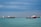 Large ships are positioned on a calm ocean under a cloudy sky. The sea appears serene with visible waves, and there is a line of blue barriers floating on the water. The ships vary in size and are distributed across the horizon, indicating a busy maritime environment.