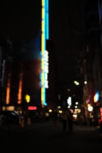 Blurred image of an urban night scene with bright neon lights in blue and yellow adorning a tall building. The street is dimly lit with soft glows from various indistinct signs and lights. The overall atmosphere is mysterious and dreamlike.