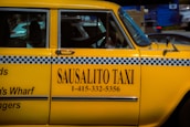 A bright yellow taxi car is prominently displayed, featuring a black and white checker pattern along its side. The words 'SAUSALITO TAXI' along with a phone number are clearly visible on the car's door. The car is parked next to other vehicles, and part of the street can be seen in the background.