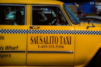 A bright yellow taxi car is prominently displayed, featuring a black and white checker pattern along its side. The words 'SAUSALITO TAXI' along with a phone number are clearly visible on the car's door. The car is parked next to other vehicles, and part of the street can be seen in the background.