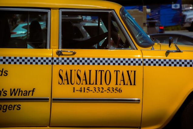 A bright yellow taxi car is prominently displayed, featuring a black and white checker pattern along its side. The words 'SAUSALITO TAXI' along with a phone number are clearly visible on the car's door. The car is parked next to other vehicles, and part of the street can be seen in the background.