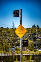 A traffic signpost with a yellow diamond-shaped sign displaying the message 'RAMP METERED WHEN FLASHING' and two traffic lights, surrounded by lush green foliage and a clear blue sky. In the background, buildings and trees can be seen.