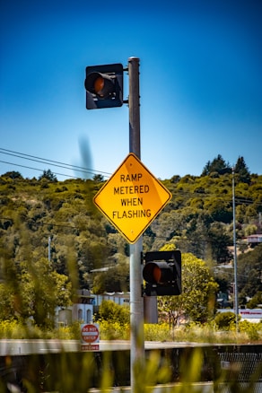 A traffic signpost with a yellow diamond-shaped sign displaying the message 'RAMP METERED WHEN FLASHING' and two traffic lights, surrounded by lush green foliage and a clear blue sky. In the background, buildings and trees can be seen.