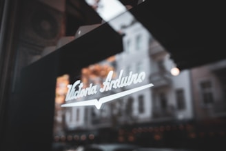 A reflection on a window glass shows the background of a vintage building with autumn trees. The words 'Victoria Arduino' are prominently displayed in stylish white lettering on the glass.