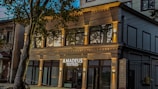 A modern building with a stylish facade features large windows and is identified by a sign that reads 'Amadeus Textiles'. The structure is illuminated by warm lights, adding an inviting glow. A tree partially obstructs the view, adding a touch of nature to the urban setting.