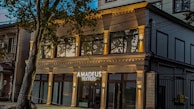 A modern building with a stylish facade features large windows and is identified by a sign that reads 'Amadeus Textiles'. The structure is illuminated by warm lights, adding an inviting glow. A tree partially obstructs the view, adding a touch of nature to the urban setting.