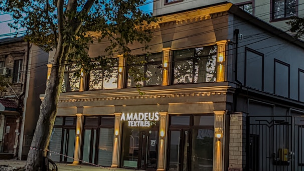 A modern building with a stylish facade features large windows and is identified by a sign that reads 'Amadeus Textiles'. The structure is illuminated by warm lights, adding an inviting glow. A tree partially obstructs the view, adding a touch of nature to the urban setting.