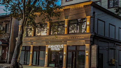 A modern building with a stylish facade features large windows and is identified by a sign that reads 'Amadeus Textiles'. The structure is illuminated by warm lights, adding an inviting glow. A tree partially obstructs the view, adding a touch of nature to the urban setting.