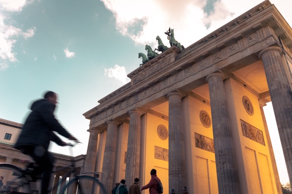 The image captures the Brandenburg Gate, a neoclassical monument in Berlin, dramatically lit from below against a partly cloudy sky. A cyclist is seen in motion in the foreground, adding dynamic movement, while several people stand near the gate, highlighting its grandeur and historical significance.