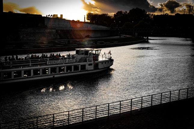 A boat passes through a tranquil river during sunset, with silhouettes of people visible on board. The sky is partially cloudy, and the sun casts a warm glow, creating reflections on the water. Trees and a bridge are visible in the background, along with a wall lining the riverbank.