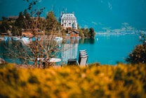 An elegant, historic-looking building sits on the shore of a serene lake, surrounded by lush greenery and distant mountains. Chairs and tables are visible in the foreground, slightly out of focus, and the vibrant turquoise water reflects the sky and surrounding landscape.