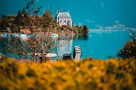 An elegant, historic-looking building sits on the shore of a serene lake, surrounded by lush greenery and distant mountains. Chairs and tables are visible in the foreground, slightly out of focus, and the vibrant turquoise water reflects the sky and surrounding landscape.