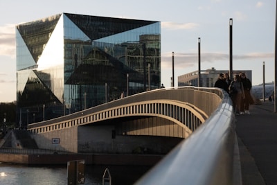 A modern, angular glass building reflects the surrounding urban landscape in the evening light. A bridge with a sleek, curved design extends toward the building, with a few people walking along it. The structure's geometric patterns and the clear sky enhance its contemporary aesthetic.