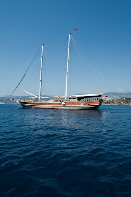 A large wooden sailing yacht is anchored on a calm body of water, with two tall masts and sails furled. The sea is a deep blue, rippling slightly under the clear, sunny sky. Near the horizon in the background, there are rolling hills and distant buildings on the coastline.