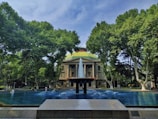 A grand building with a dome sits elegantly behind a water fountain surrounded by tall, lush green trees. The fountain features multiple jets with water gracefully arching. Engravings are visible on a stone plaque in the foreground.