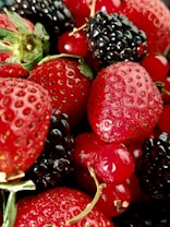 A close-up of a variety of fresh berries, prominently featuring strawberries, blackberries, and red currants. The berries display a vibrant, glossy texture, indicating freshness and juiciness. The green leaves of the strawberries add a contrasting pop of color.