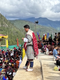 A man dressed in traditional attire stands on the edge of a platform, overlooking a large crowd seated on the ground. The background reveals a scenic view of green mountains under a cloudy sky. Colorful flags are visible throughout the crowd, suggesting a cultural or festive event.
