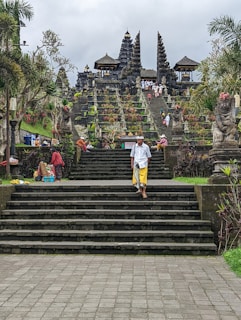 Lush green scenery surrounds a flight of stone steps leading up to a grand temple structure with multiple spires and shrines, adorned with intricate carvings. The steps are populated with several people, some wearing traditional attire, and there are small stalls on the sides selling goods. Palm trees and other tropical plants add to the vibrant setting.