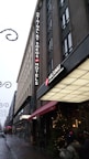 A tall hotel building with a sign reading 'ORIGINAL BY SOKOS HOTELS' and another sign displaying 'HELSINKI'. The street is adorned with festive lights and decorations, suggesting a holiday atmosphere. There is a Christmas tree with lights near the entrance, and several people are visible on the sidewalk.