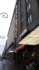 A tall hotel building with a sign reading 'ORIGINAL BY SOKOS HOTELS' and another sign displaying 'HELSINKI'. The street is adorned with festive lights and decorations, suggesting a holiday atmosphere. There is a Christmas tree with lights near the entrance, and several people are visible on the sidewalk.